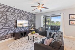 Un salon moderne avec un mur orné d’un motif géométrique, un ventilateur de plafond, un ensemble de canapés, une table basse et un téléviseur.