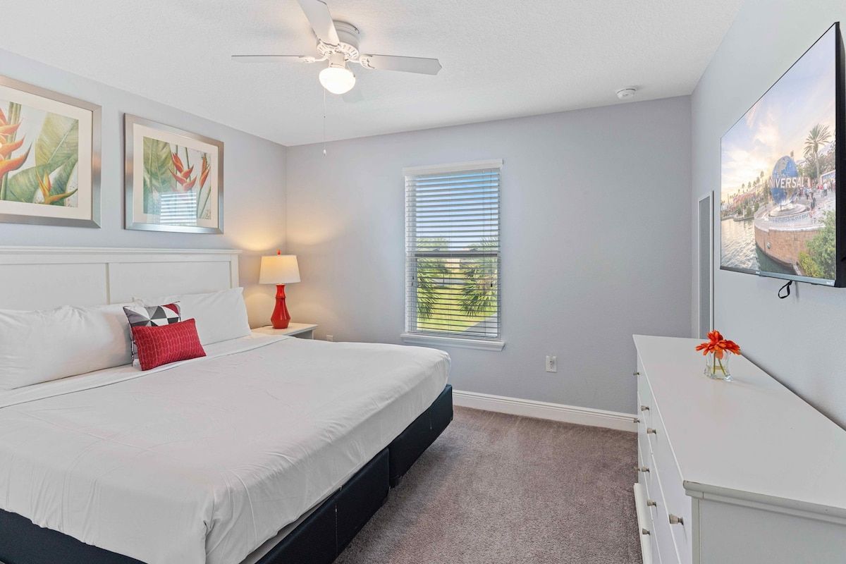 A bedroom with a bed, a nightstand, a lamp, a ceiling fan, a window with blinds, and a wall-mounted TV.