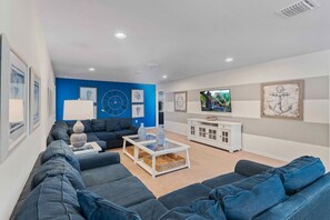 A modern living room with a blue accent wall, a large sofa set, a coffee table, and a television. - Lake View! 8BR Home w Private Pool Spa near Disney (Kissimmee)
