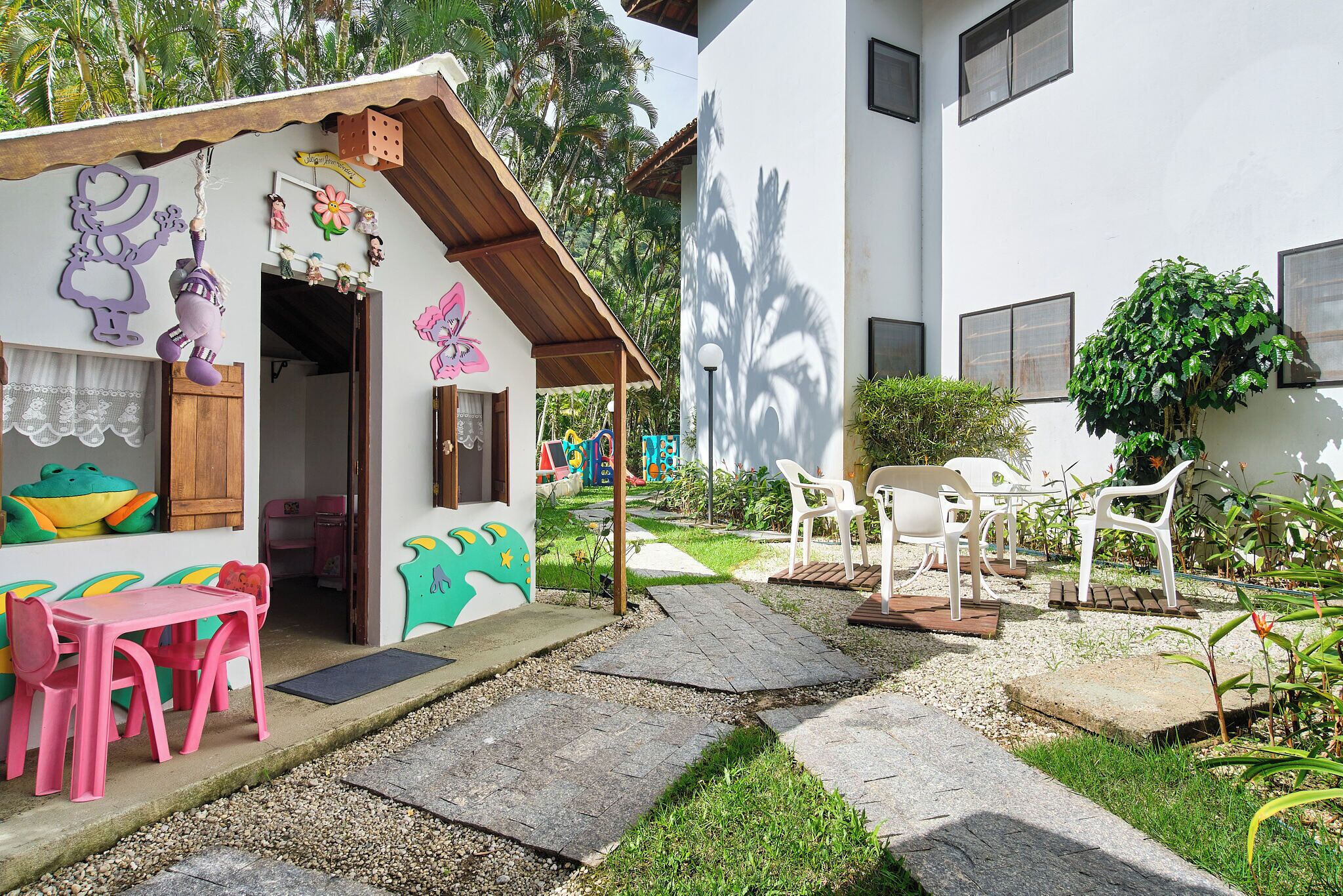 A small, colorful building with a play area, including a pink table and chairs, and a garden with white plastic chairs.