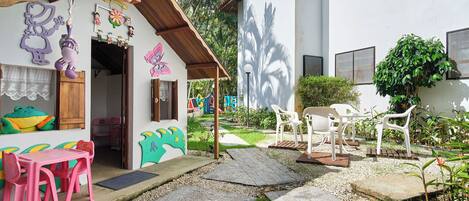 A small, colorful building with a play area, including a pink table and chairs, and a garden with white plastic chairs.
