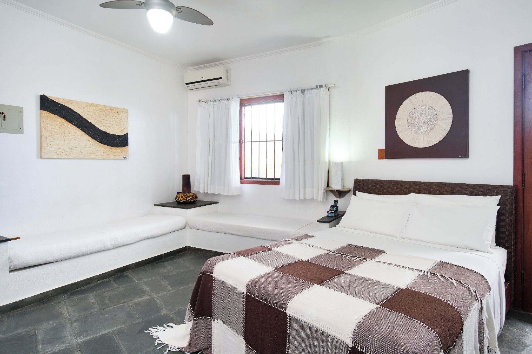 Un dormitorio con cama, mesita de noche, lámpara, ventana con cortinas y obras de arte en las paredes.