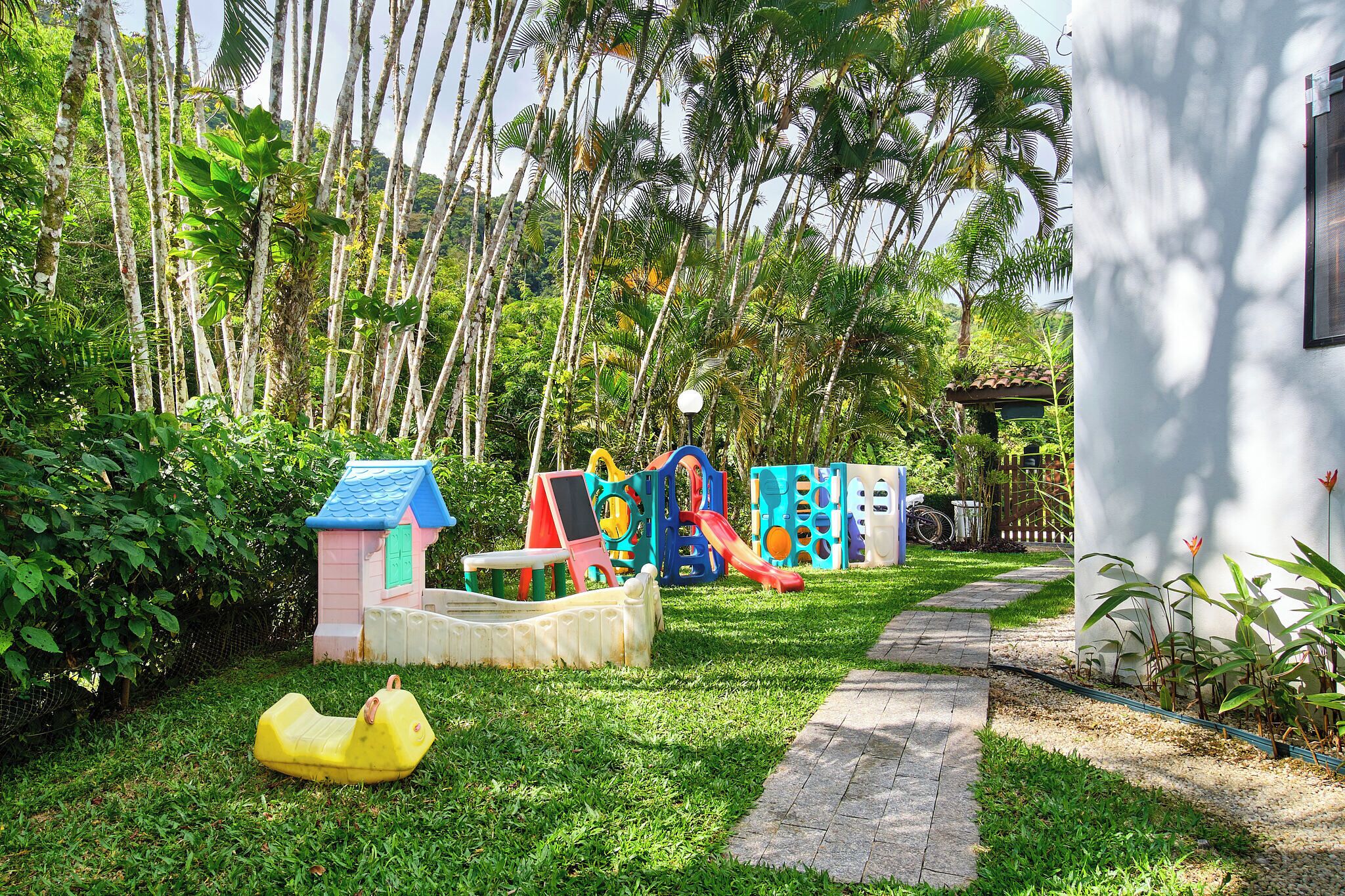 A playground with slides and a small house structure, surrounded by lush greenery and tall trees.