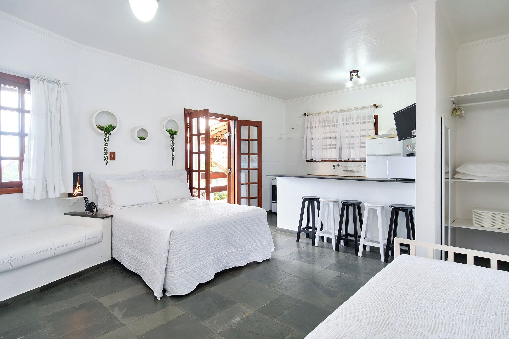 A white-themed bedroom with a bed, a bench, a desk with a computer, and a small kitchen area.