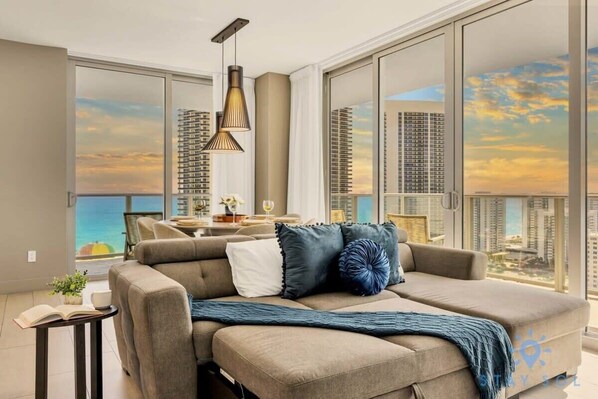 Un salon moderne avec un grand canapé, une table à manger et une vue sur de hauts immeubles et un coucher de soleil.