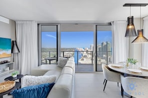 A modern living room with a sofa, dining table, and chairs, a large window with a city view, and pendant lighting. - Luxury 2Br| Front Ocean| Bay View| Amazing Pools (Hollywood)