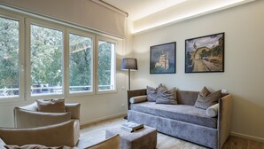 Una sala de estar moderna con un sofá gris, un sillón a juego y un pequeño otomaní. Hay dos cuadros enmarcados en la pared, una lámpara de pie y ventanas amplias con vistas a árboles.