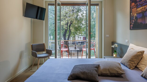Un dormitorio con cama, almohadas, televisor, una silla y un balcón con sillas rojas y una mesa.