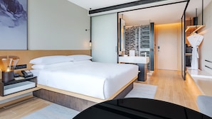 A modern hotel room with a large bed, bedside table with a lamp, a desk, and a chair. There is a view of the bathroom and a wardrobe.