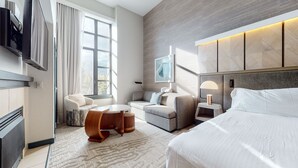 Una habitación de hotel moderna con una cama grande, un sofá, una pequeña mesa y un escritorio. Hay una ventana con cortinas, un cuadro en la pared y una lámpara de techo.