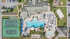 Vista aérea de un complejo turístico con una gran piscina, canchas de tenis y una zona de playa.