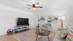 Un salon moderne doté d’un téléviseur à écran plat, d’un ventilateur de plafond, d’un canapé, d’une table basse et de deux tableaux accrochés au mur.