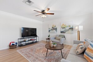 A modern living room with a flat-screen TV, a ceiling fan, a sofa, a coffee table, and two paintings on the wall. - Beach Bungalow- Perfect duplex for larger families (Fort Myers Beach)