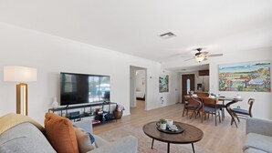 Un salon moderne avec un téléviseur à écran plat, un coin repas avec des chaises en bois et une cuisine en arrière-plan.