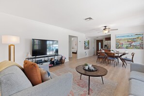 A modern living room with a flat-screen TV, a dining area with wooden chairs, and a kitchen in the background. - Beach Bungalow- Perfect duplex for larger families (Fort Myers Beach)