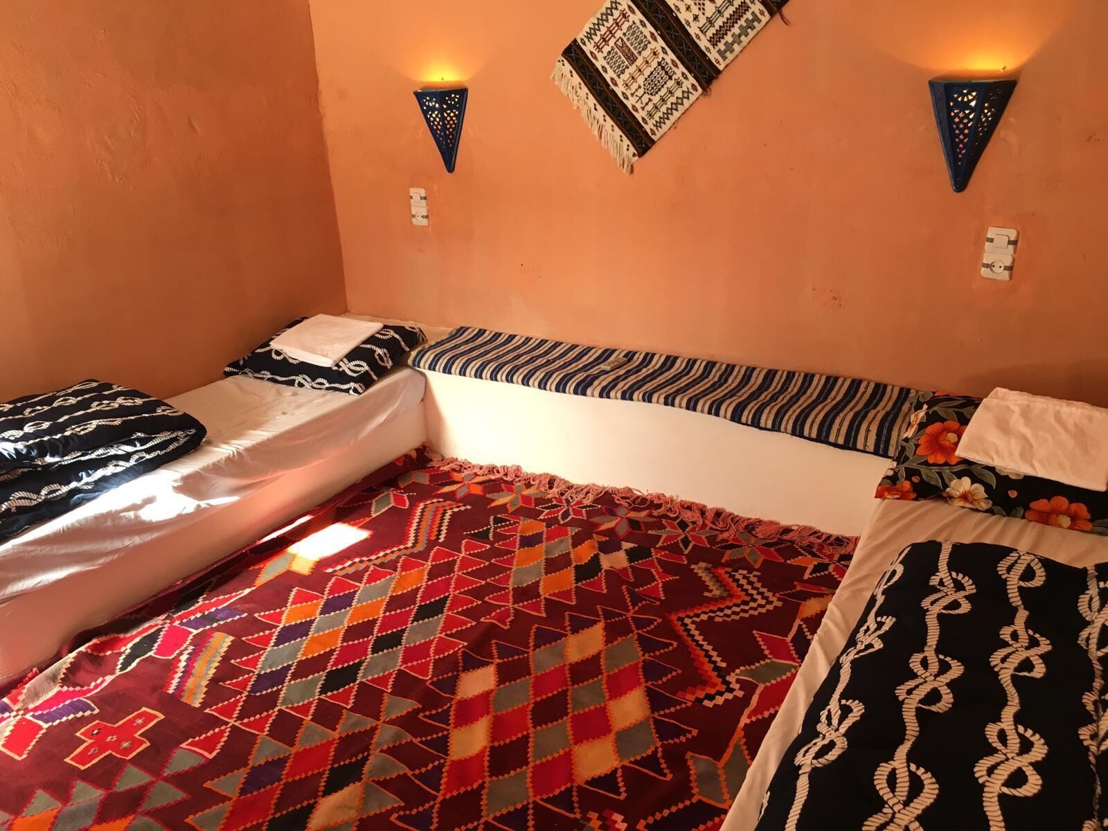 A room with two beds, each with patterned bedding, a colorful geometric rug, and wall-mounted lights.