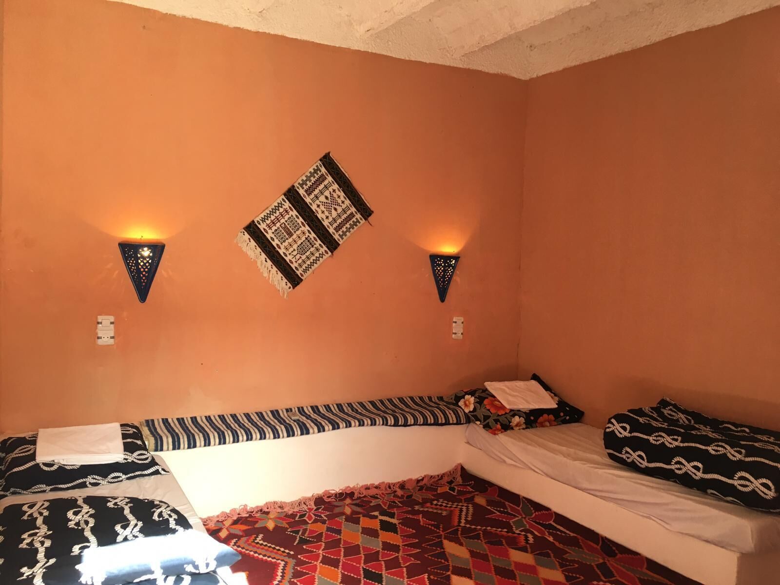 A room with two beds, each with patterned bedding. There are wall-mounted lights and a decorative wall hanging.