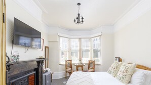 Une chambre d’hôtel avec une cheminée, une télévision, une grande fenêtre avec des stores, un lit agrémenté d’oreillers à motifs floraux et un lustre.