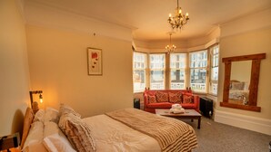 A bedroom with a large bed, a red sofa, a wooden coffee table, and a bay window with blinds. - Pittodrie Guest House - Room 2 (Brighton)