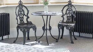 A room with a round table, two ornate chairs, a chandelier, and a window with blinds.