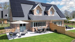 A two-story house with a sloped roof, solar panels, a patio with outdoor furniture, and a brick exterior. - Moderne Doppelhaushälfte in Niebüll (Niebüll)