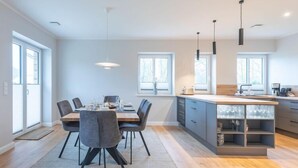 A modern kitchen with a dining area, large windows, and a central island with a built-in sink. - Moderne Doppelhaushälfte in Niebüll (Niebüll)