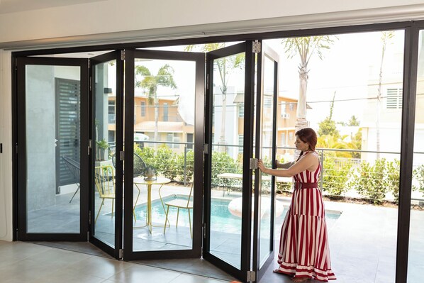 Una mujer con un vestido rojo y blanco a rayas abre un ventanal corredero hacia un balcón con piscina.