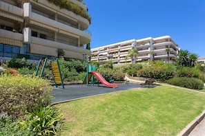 A playground with slides and a bench, surrounded by greenery and a building with balconies.