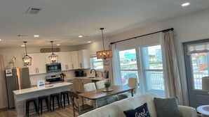 A modern kitchen with white cabinets, a central island, and bar stools. There is a dining area with a wooden table and chairs, and a living room with a sofa and decorative pillows. - Beautiful Beach Home Steps from the crystal blue waters of Navarre Beach! (Navarre)