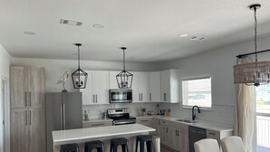 A modern kitchen with white cabinets, a stainless steel refrigerator, a microwave, and a large island with bar stools. - Beautiful Beach Home Steps from the crystal blue waters of Navarre Beach! (Navarre)
