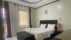 A bedroom with a bed, a nightstand, a lamp, a window with curtains, and a wall-mounted artwork.