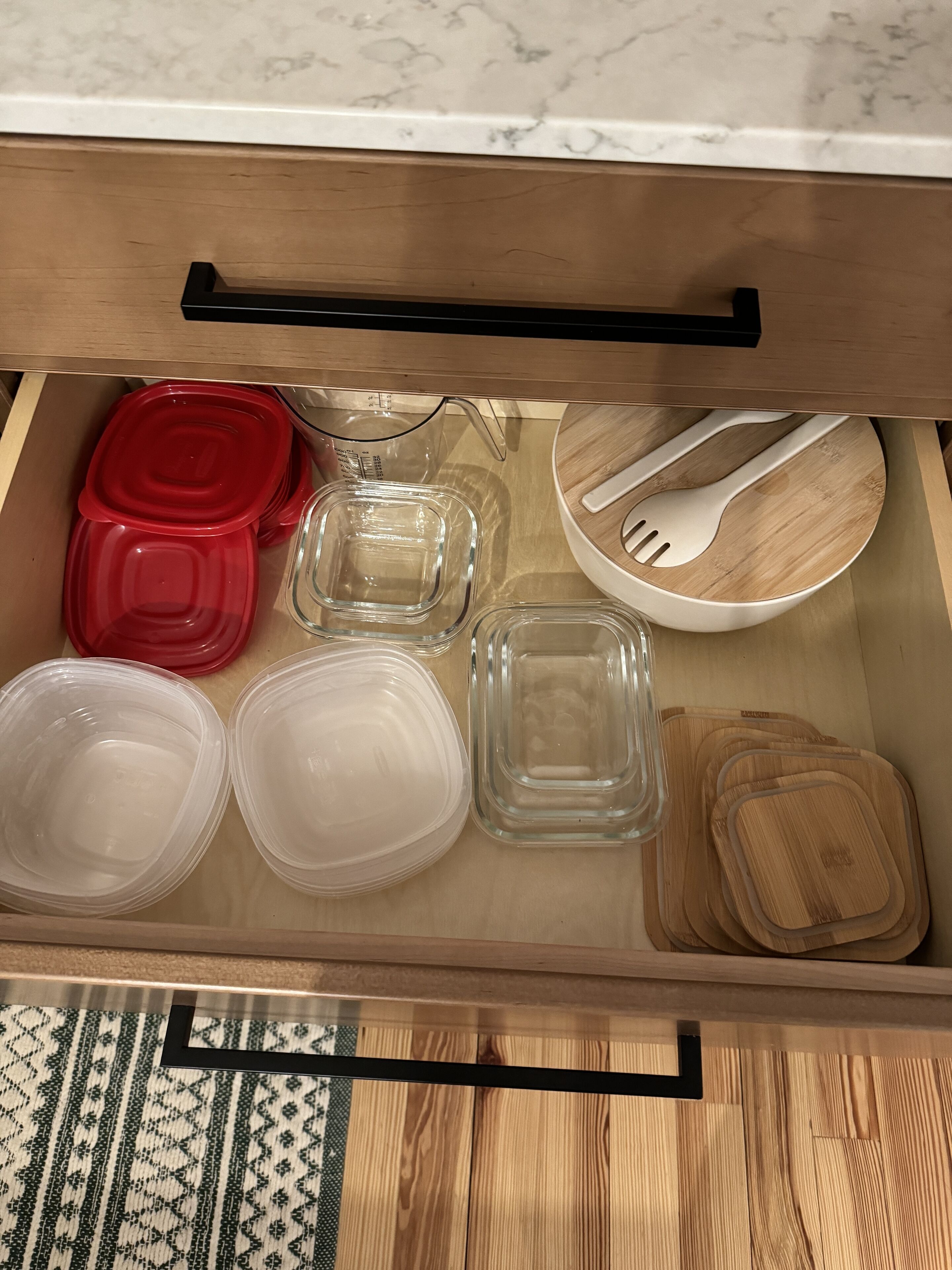 A kitchen drawer with various containers, including red and clear plastic containers, a glass measuring cup, and wooden cutting boards.