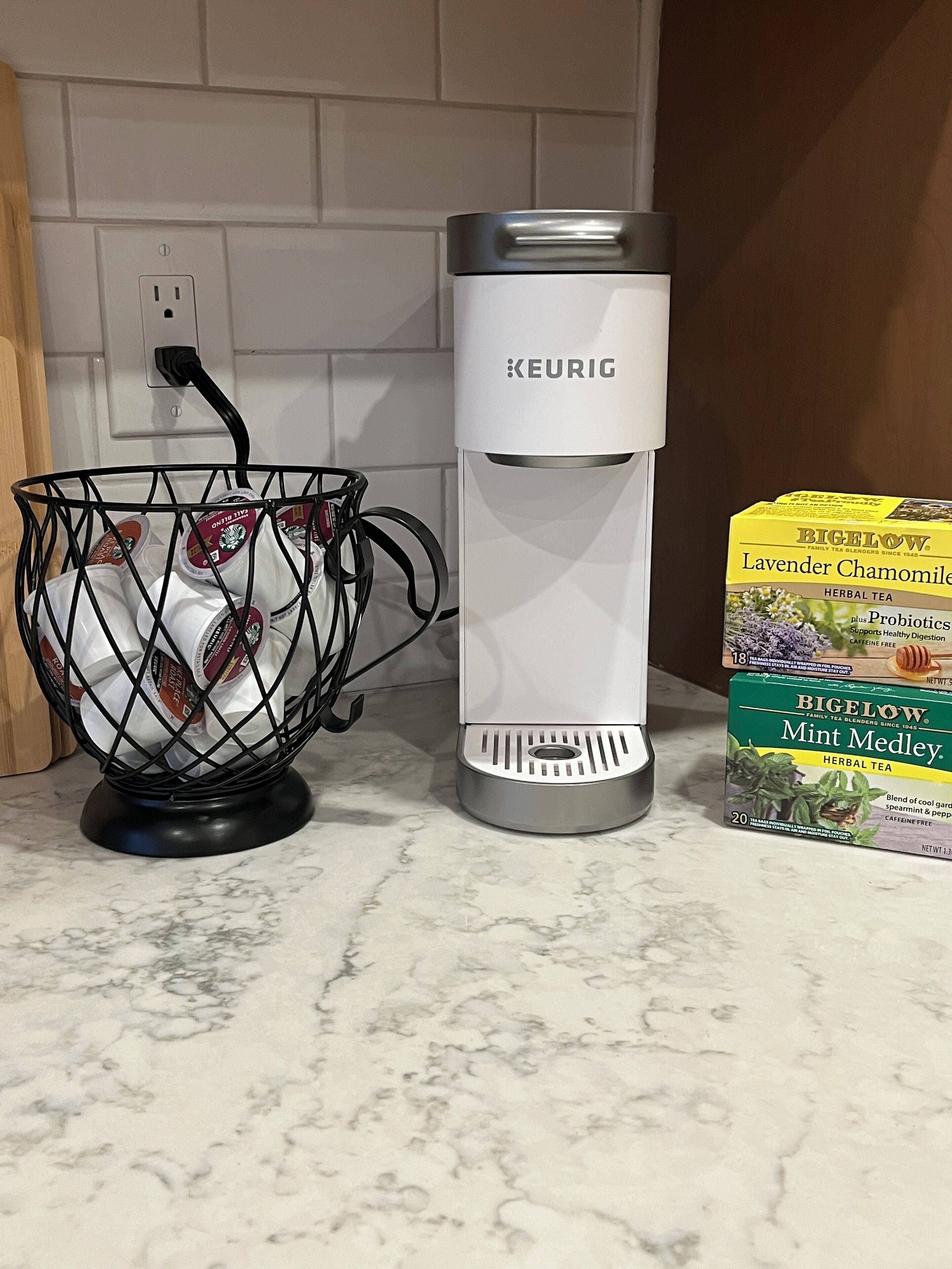Une machine à café Keurig posée sur un comptoir de cuisine, avec un panier de dosettes de café et deux boîtes de thé aux plantes de la marque Bigelow.