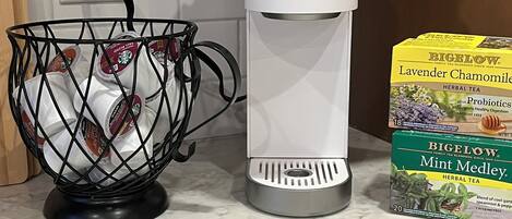 Une machine à café Keurig posée sur un comptoir de cuisine, avec un panier de dosettes de café et deux boîtes de thé aux plantes de la marque Bigelow.