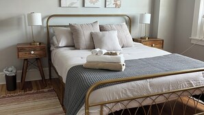 A neatly arranged bedroom with a metal bed frame, a grey patterned bedspread, and a wooden floor. There are two bedside tables with lamps and a trash bin.