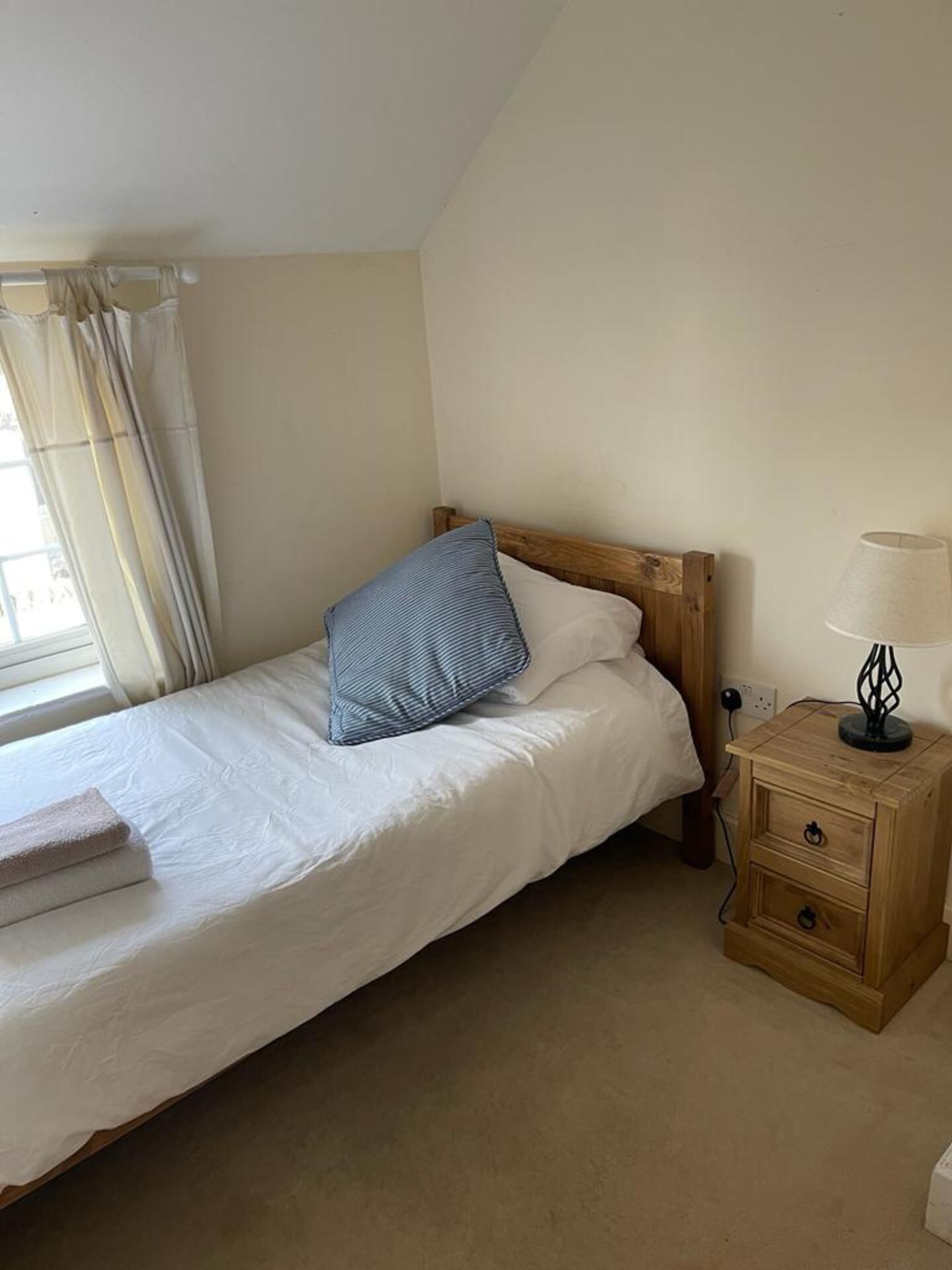A single bed with white bedding, a wooden headboard, a blue pillow, and a bedside table with a lamp.