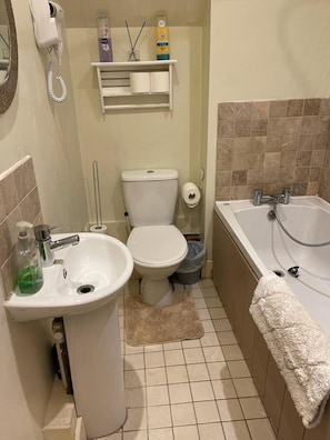 A compact bathroom with a toilet, sink, and bathtub. There is a shelf above the toilet holding toiletries and a towel hanging on the wall.