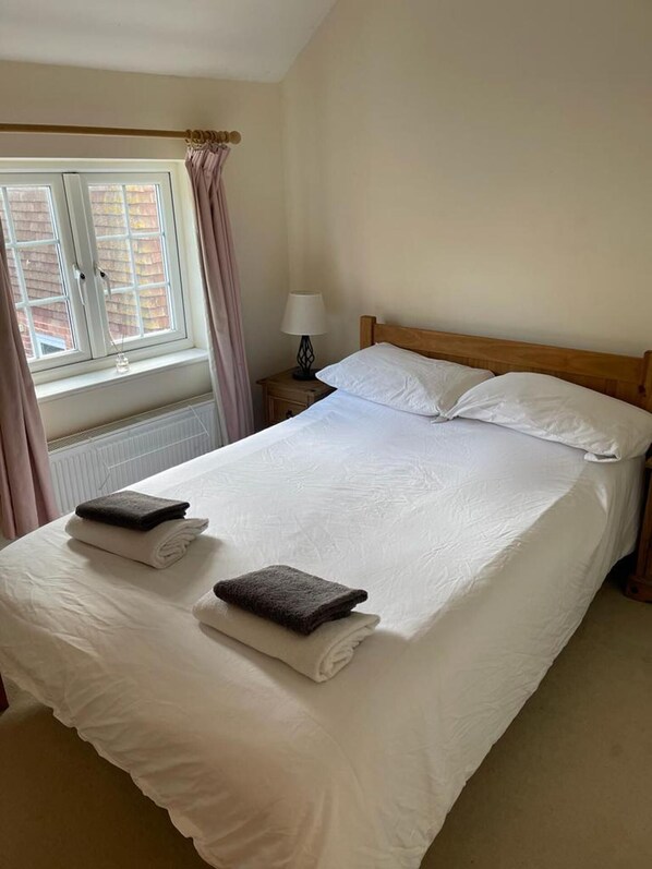 A neatly made bed with white linens and two folded towels, a bedside lamp, and a window with curtains.