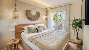 A modern bedroom with a wooden headboard, a large bed with multiple pillows, a small wooden stool, a hanging light fixture, and a view of the outdoors through a sliding door.