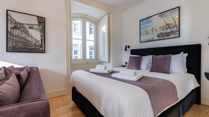 A hotel room with a large bed, a view of the street through an arched window, and a painting of a coastal scene.