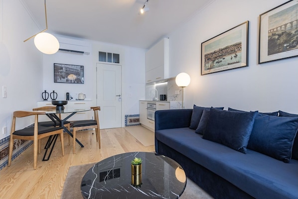 A modern living room with a blue sofa, a dining table with chairs, a kitchen area, and artwork on the walls.