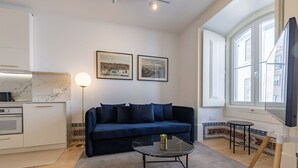 Una sala de estar moderna con un sofá azul oscuro, una mesa de centro de vidrio y una cocina con armarios blancos y salpicadero de mármol.