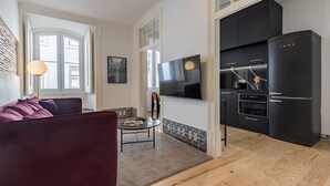 A modern living room with a sofa, a coffee table, and a TV. There is a kitchen area with a black refrigerator and a dining table. - Figueira Quality III by LovelyStay (Lisboa)