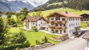 A multi-story chalet with balconies, surrounded by lush greenery and mountains in the background.