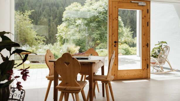 A wooden dining table with chairs, a rocking chair, and a view of a forest through a glass door.