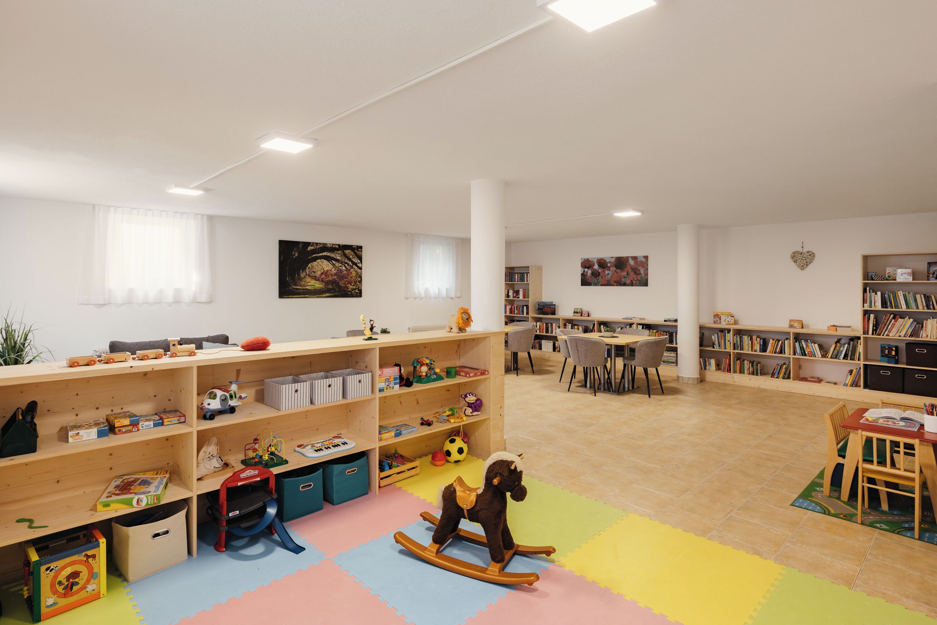 Ein geräumiger Raum mit einem großen, hölzernen Bücherregal voller Bücher und Spielzeug, einem kleinen Tisch mit Stühlen und einer bunten Spieledecke auf dem Boden.