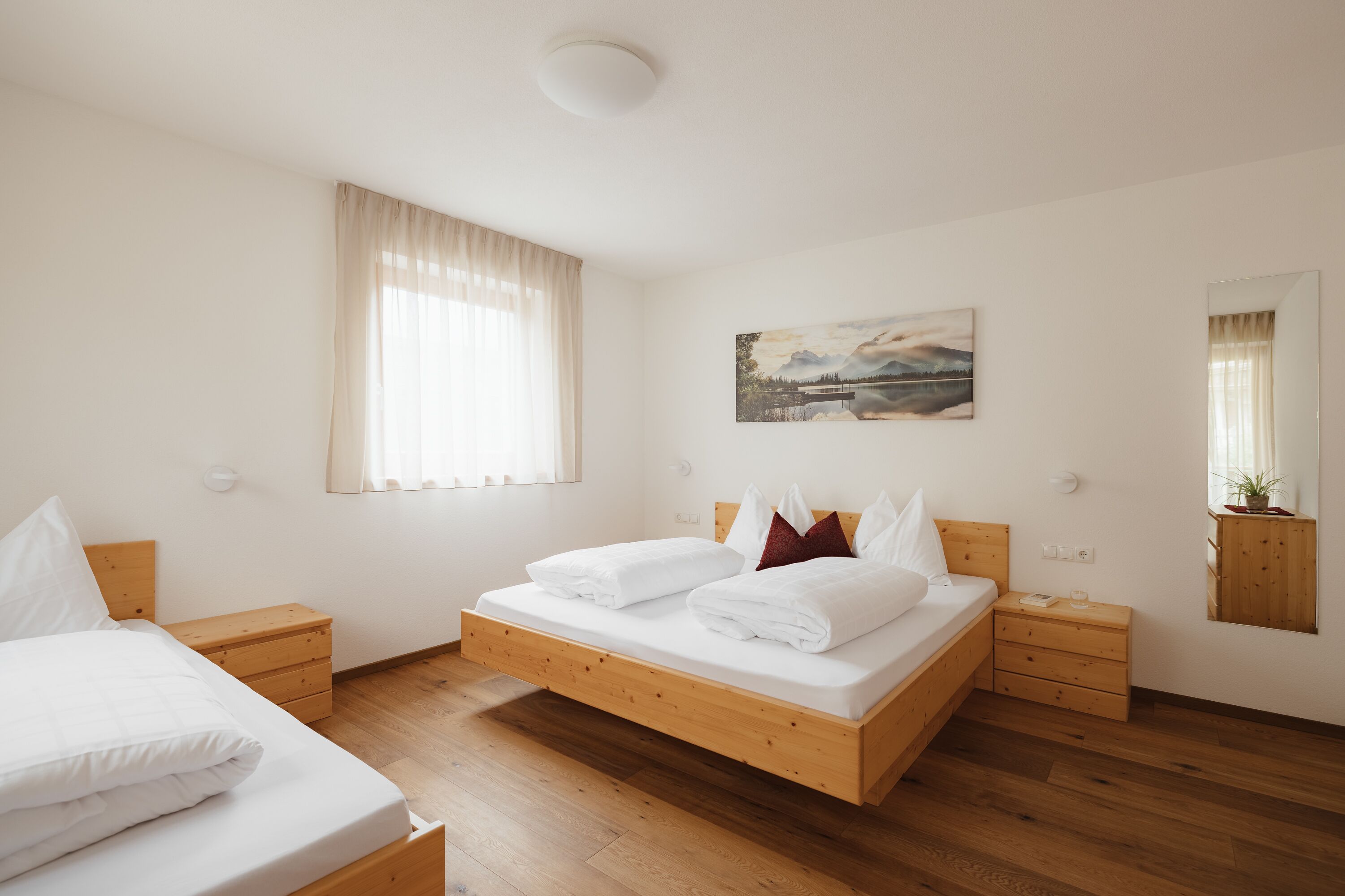 Ein Schlafzimmer mit einem hölzernen Bett, zwei Nachttischen, einem Fenster mit Vorhängen, einem Gemälde an der Wand und einem Spiegel.