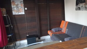 Ein Zimmer mit Holzboden, einem grauen Sofa mit orangefarbenen und grauen Kissen, einem kleinen Holztisch, einem roten Vorhang und einem Wandbild von einem Leoparden.