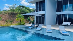 Un espace piscine moderne avec une eau d’un bleu limpide, quatre chaises longues et un grand parasol.