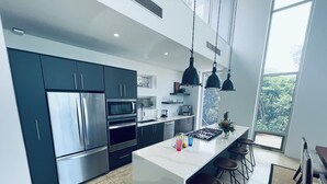 Une cuisine moderne dotée d’appareils électroménagers en acier inoxydable, d’un îlot central avec des tabourets de bar et de grandes fenêtres offrant une vue sur la verdure.